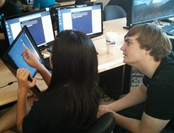 A college student holds a laptop diagonally in the air, while a second student looks tries to work through the puzzle on the screen.